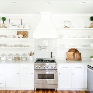 May include: A bright, modern kitchen with white cabinetry, countertops, and shelving. Stainless steel range and hood. Decorative items include small potted plants, framed art, and wooden cutting boards. The kitchen has a clean, minimalist aesthetic.