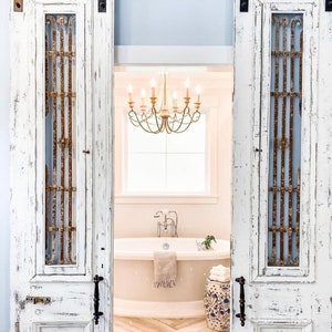 May include: White distressed barn doors with decorative metal grates open to reveal a bathroom. A chandelier hangs above a bathtub, with a patterned stool and herringbone floor.