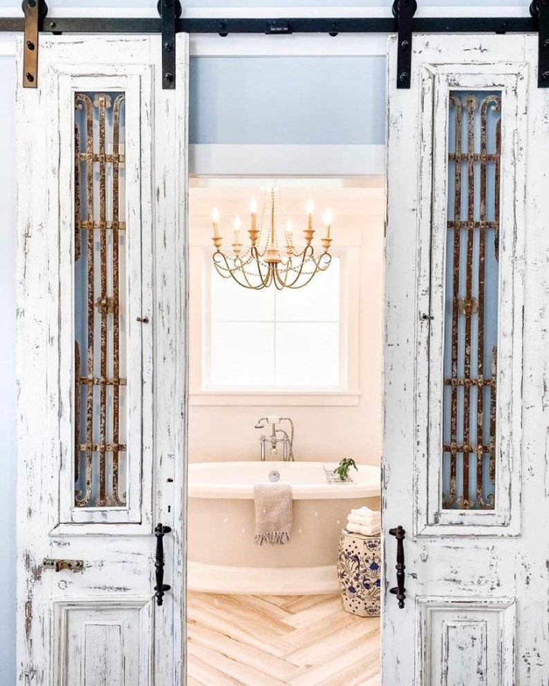 May include: White distressed barn doors with decorative metal grates open to reveal a bathroom. A chandelier hangs above a bathtub, with a patterned stool and herringbone floor.