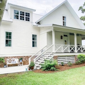May include: A two-story white house with a porch, stairs, and a metal roof. The house has multiple windows and is surrounded by green grass and landscaping. A white bench with blue and white striped pillows sits on the porch.