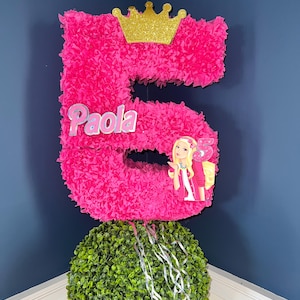 May include: A pink number 5 piñata with a gold crown on top. The piñata has the name "Paola" written on it and a Barbie doll image on the side. The piñata is sitting on top of a green bush.