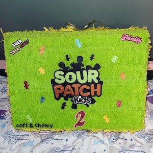 May include: A green Sour Patch Kids piñata with the text "Sour Patch Kids" and "soft & chewy" on it. The piñata is decorated with colorful gummy candies and the number "2" in pink glitter. The piñata is on a white and blue patterned surface.
