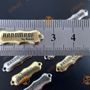 Könnte beinhalten: Ein goldenes Metalletikett mit den Worten "HANDMADE by Susan" eingraviert. Das Etikett ist etwa 3,5 cm lang und hat zwei Löcher zum Anbringen an einem Produkt.