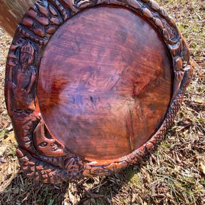 Puede incluir: Una bandeja redonda de madera de color marrón oscuro con un centro pulido y liso. El borde elevado presenta intrincadas figuras talladas a mano, incluyendo una persona, varios animales y patrones texturizados que rodean todo el perímetro.