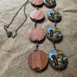 Puede incluir: Un collar con una cadena de tono bronce conectada a cuatro discos de madera circulares y cuatro colgantes de resina circulares. Cada colgante de resina muestra un elefante en un paisaje con el texto ORULA impreso en letras doradas.