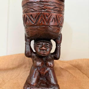 Puede incluir: Una escultura de madera tallada a mano en color marrón oscuro que representa una figura arrodillada sosteniendo un gran recipiente cilíndrico con tapa sobre su cabeza. El recipiente presenta bandas texturizadas y grabados en zigzag.