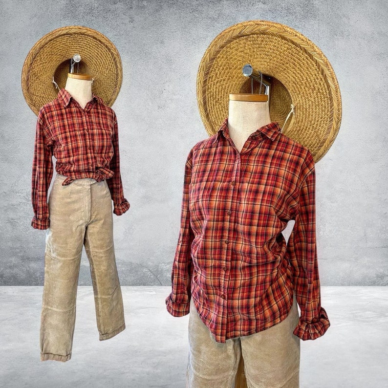 K&ouml;nnte beinhalten: Ein rotes und oranges kariertes Flanellhemd mit langen &Auml;rmeln und Knopfleiste. Das Hemd wird von einem Mannequin mit einem Strohhut und einer beigefarbenen Cordhose getragen.