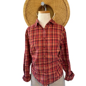 K&ouml;nnte beinhalten: Ein lang&auml;rmliges Hemd mit rotem und schwarzem Karomuster und Knopfleiste. Das Hemd befindet sich auf einem Mannequin, das beige Shorts und einen Strohhut tr&auml;gt.