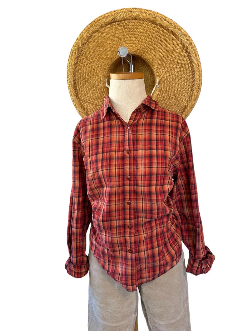 K&ouml;nnte beinhalten: Ein lang&auml;rmliges Hemd mit rotem und schwarzem Karomuster und Knopfleiste. Das Hemd befindet sich auf einem Mannequin, das beige Shorts und einen Strohhut tr&auml;gt.