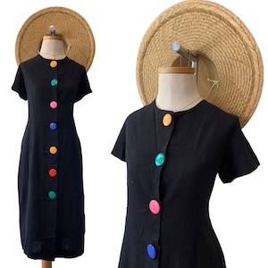 Op de afbeelding: Twee zwarte, korte mouwen jurken met kleurrijke knopen aan de voorkant. De jurk aan de linkerkant heeft een ronde hals en de jurk aan de rechterkant heeft een ronde hals.