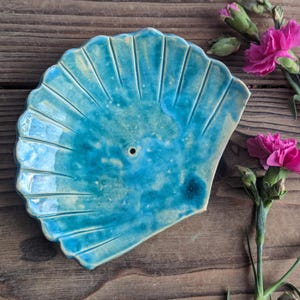 Peut inclure: Un brûleur d'encens en forme de coquille de mer en céramique bleu sarcelle avec un petit trou au centre. Le brûleur est posé sur une surface en bois.