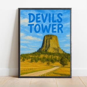 Może przedstawiać: Wydruk w ramie przedstawiający Devils Tower National Monument w Wyoming. Obraz przedstawia dużą formację skalną wznoszącą się z trawiastego pola z drogą gruntową prowadzącą do jej podstawy. Niebo jest niebieskie z białymi chmurami. Tekst "Devils Tower" jest napisany niebieskimi literami u góry wydruku.