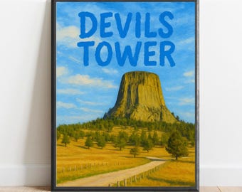 May include: A framed print of Devils Tower National Monument in Wyoming. The image shows a large rock formation rising from a grassy field with a dirt road leading to the base. The sky is blue with white clouds. The text "Devils Tower" is written in blue letters at the top of the print.