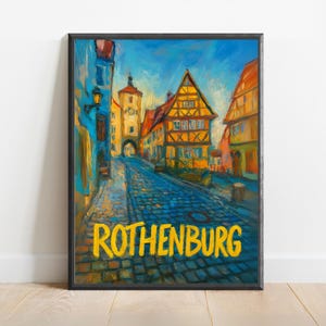 May include: A framed art print showing a European street scene. The artwork depicts a cobblestone street lined with colourful buildings under a blue sky. The word "ROTHENBURG" is printed in yellow at the bottom of the image.