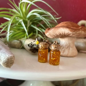 Puede incluir: Un par de pendientes dorados con pequeños frascos de vidrio llenos de purpurina dorada. Los frascos tienen tapas de corcho y están unidos a ganchos dorados.