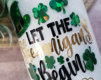 Puede incluir: Un vaso blanco con purpurina verde y dorada. El vaso tiene un diseño negro y dorado que dice "Let the Shenanigans Begin". Hay diseños de trébol verde en el vaso.
