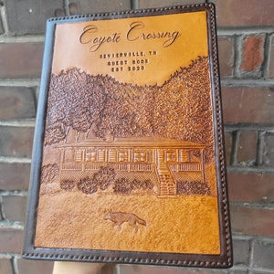 May include: A brown leather guest book with the title "Coyote Crossing" embossed on the front. The book also includes the text "Sevierville, TN Guest Book Est 2023". The cover features an embossed image of a cabin with a porch and a coyote.