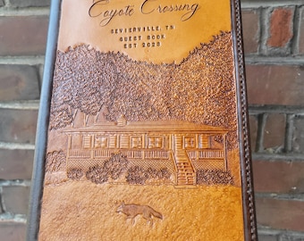 Puede incluir: Un libro de visitas de cuero marrón con el título "Coyote Crossing" en relieve en la portada. El libro también incluye el texto "Sevierville, TN Guest Book Est 2023". La portada presenta una imagen en relieve de una cabaña con un porche y un coyote.