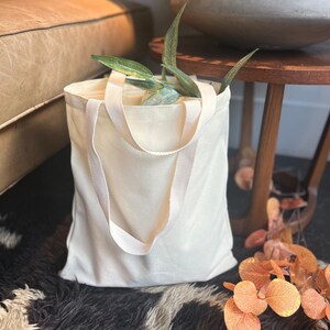 May include: A white canvas tote bag with long straps. The bag is sitting on a fuzzy rug with a potted plant and a wooden table in the background.