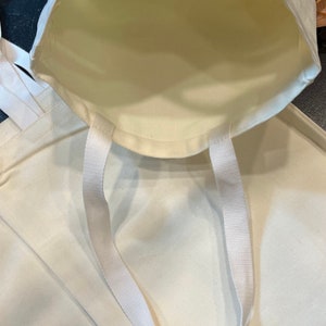 May include: Two white canvas tote bags with white straps. The bags are empty and the straps are folded over the top of the bags.