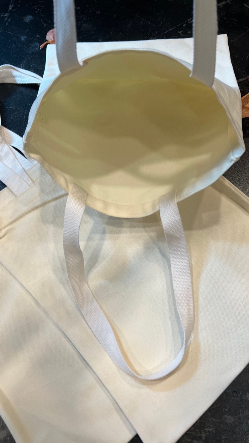 May include: Two white canvas tote bags with white straps. The bags are empty and the straps are folded over the top of the bags.