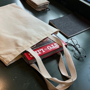 May include: A white canvas tote bag with a red book titled "Eto Life" inside. The book is about healthy and delicious ketogenic recipes. The bag is on a green surface with a pair of glasses and a black notebook.