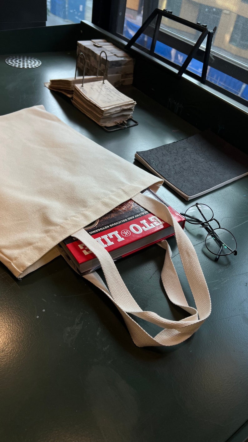 May include: A white canvas tote bag with a red book titled "Eto Life" inside. The book is about healthy and delicious ketogenic recipes. The bag is on a green surface with a pair of glasses and a black notebook.