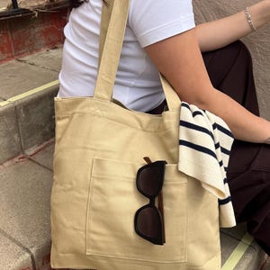 Puede incluir: Bolso tote de lona beige con un bolsillo delantero, que contiene gafas de sol negras. Un suéter a rayas está sobre el bolso. El bolso tiene correas largas. El fondo incluye una camiseta blanca y pantalones marrones.