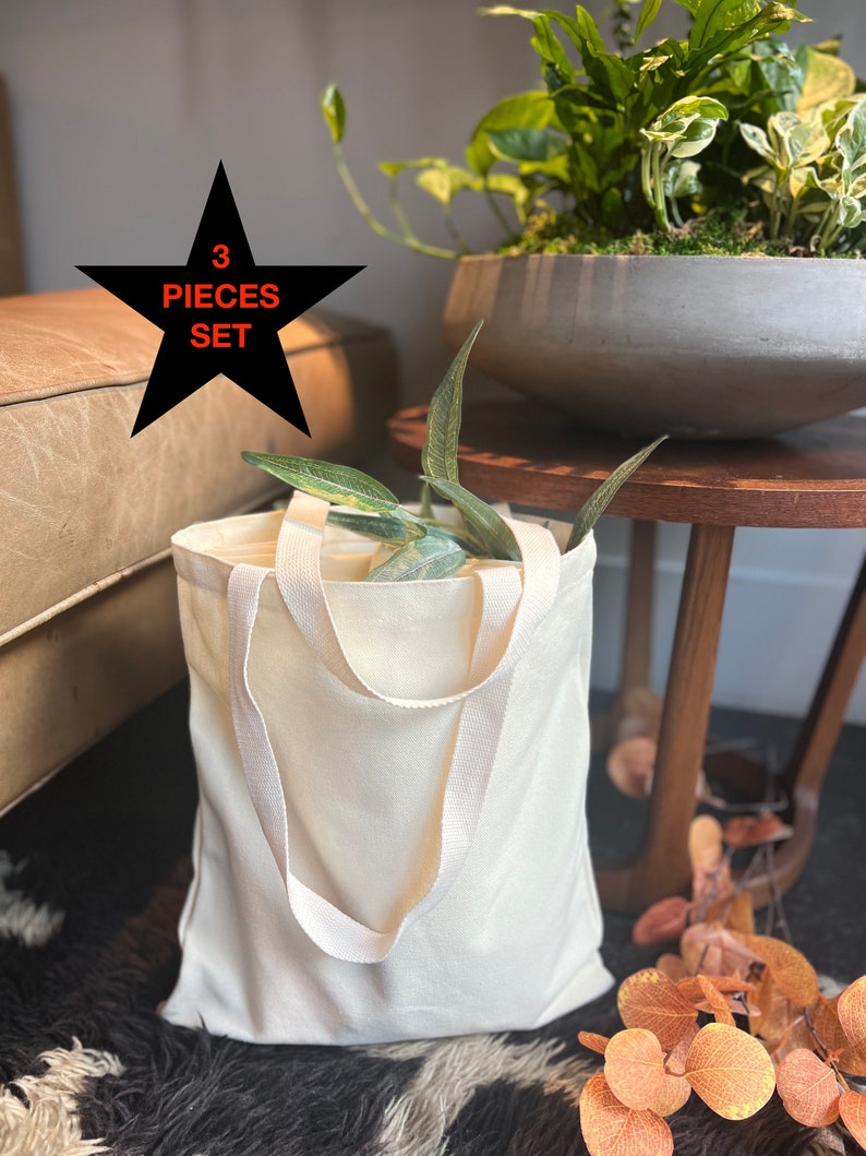 May include: A set of three off-white canvas tote bags with black handles. The bags are sitting on a brown and white fuzzy rug. A black star with the text "3 PIECES SET" is in the background.