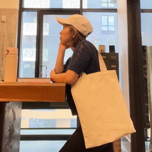 May include: A person wearing a white baseball cap and a blue sweater is holding a large, white canvas tote bag. The bag is hanging from their shoulder and is empty.
