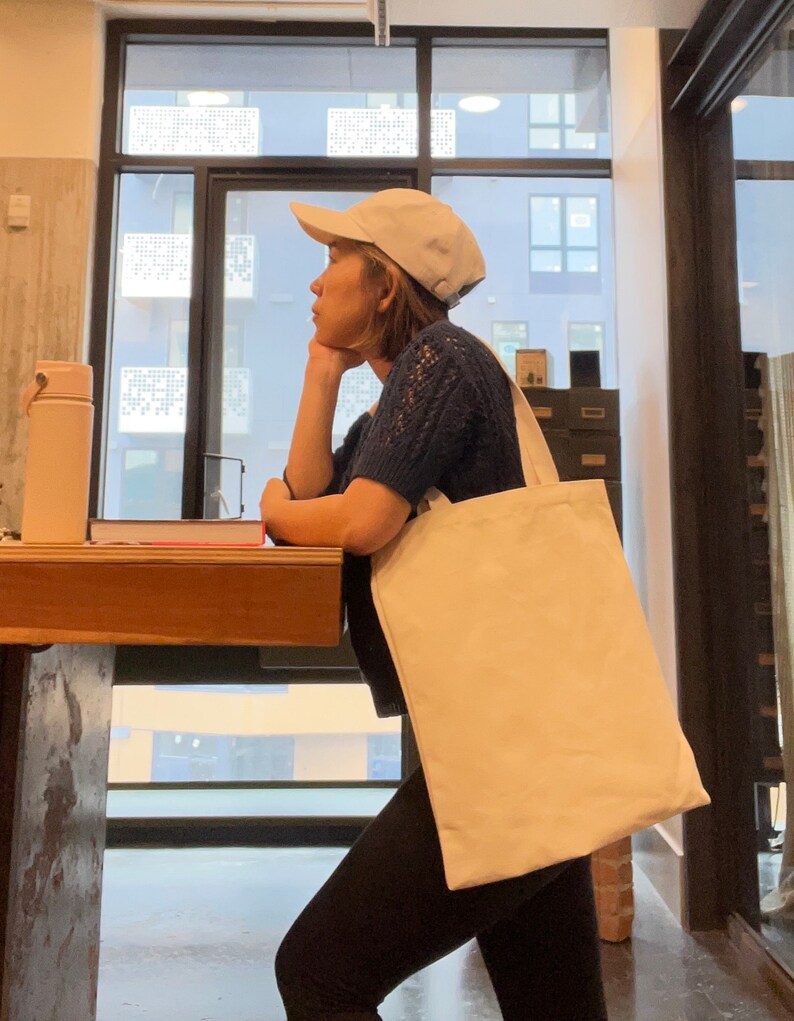 May include: A person wearing a white baseball cap and a blue sweater is holding a large, white canvas tote bag. The bag is hanging from their shoulder and is empty.