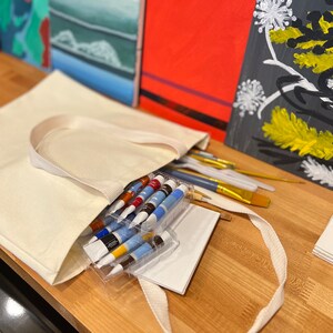 May include: A white canvas tote bag filled with art supplies, including paint tubes, brushes, and a white pad of paper. The bag is sitting on a wooden table next to three paintings.