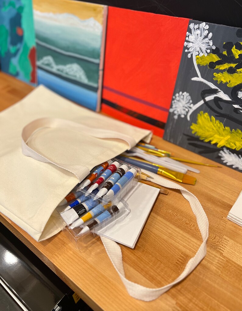 May include: A white canvas tote bag filled with art supplies, including paint tubes, brushes, and a white pad of paper. The bag is sitting on a wooden table next to three paintings.