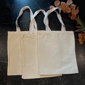 May include: Three plain white canvas tote bags with handles. The bags are stacked on top of each other.