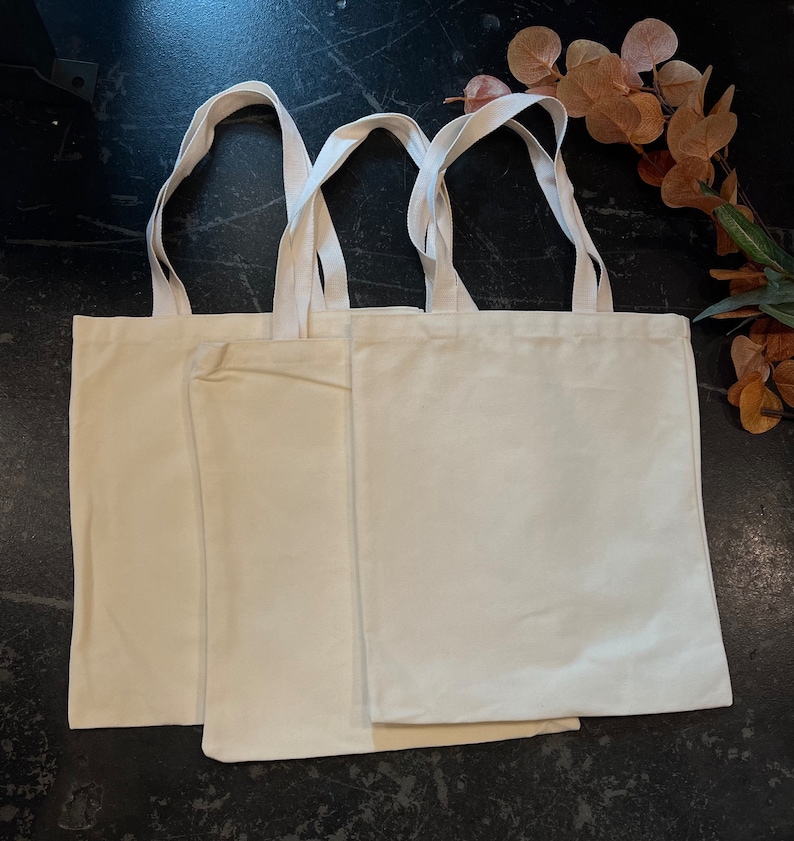 May include: Three plain white canvas tote bags with handles. The bags are stacked on top of each other.