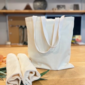 May include: Two white cotton tote bags with white handles. The bags are folded and placed on a wooden surface.