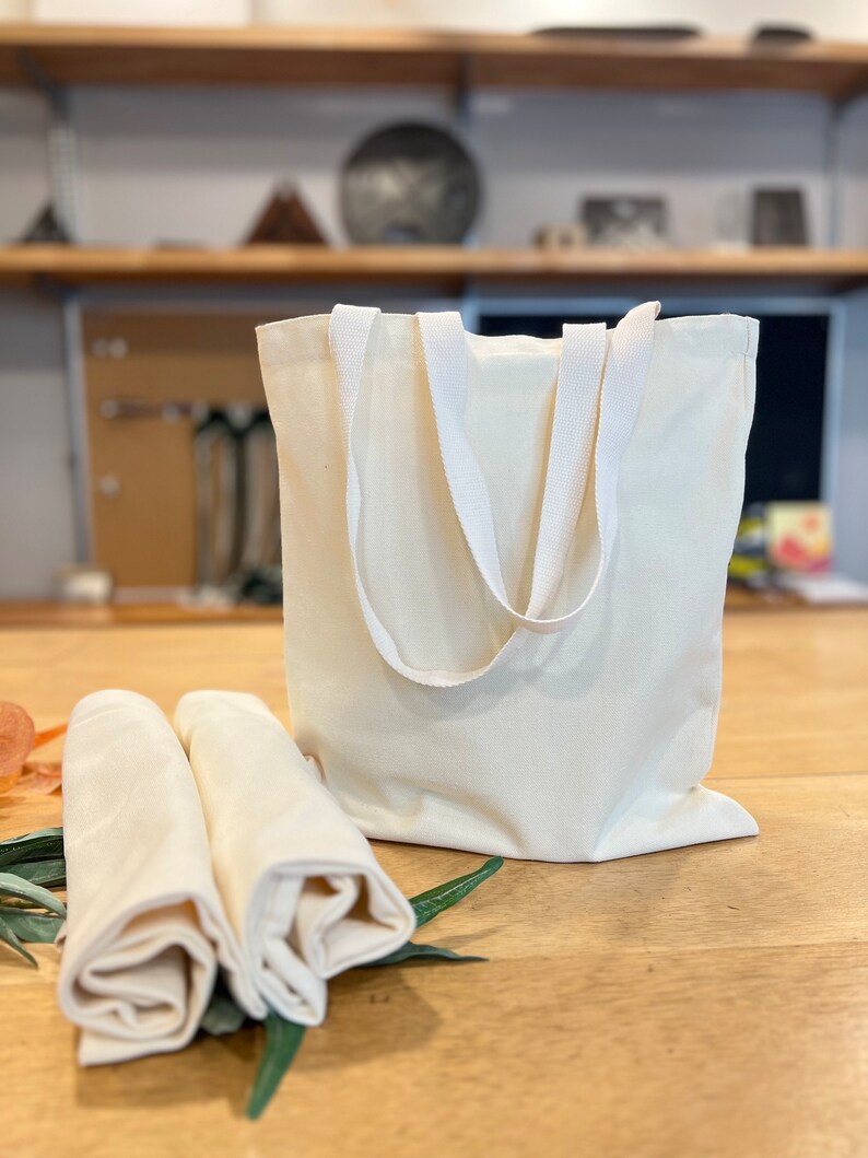 May include: Two white cotton tote bags with white handles. The bags are folded and placed on a wooden surface.