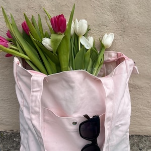 Könnte beinhalten: Eine hellrosa Canvas-Tragetasche gefüllt mit frischen Tulpen. Die Tasche hat eine Vordertasche und einen Reißverschluss. Schwarze Sonnenbrillen liegen in der Tasche. Die Tulpen sind eine Mischung aus Weiß und Dunkelrosa.