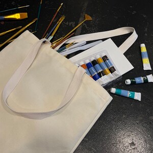 May include: A white canvas tote bag filled with art supplies, including paintbrushes, paint tubes, and a blank canvas. The bag has a white strap and is lying on a black surface.
