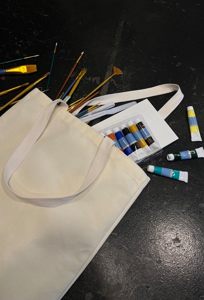 May include: A white canvas tote bag filled with art supplies, including paintbrushes, paint tubes, and a blank canvas. The bag has a white strap and is lying on a black surface.