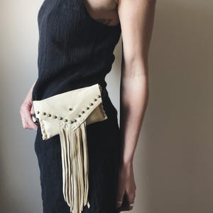 May include: A black dress with spaghetti straps. A cream-colored leather fanny pack with metal stud accents and fringe hangs at the waist. The fanny pack has a flap closure.
