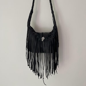 May include: Black leather shoulder bag with long fringe detailing. The bag features a braided strap and a decorative silver-toned metal accent. The bag is hanging against a plain white background.