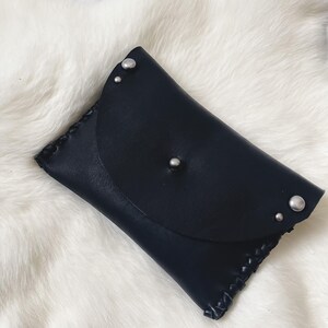 May include: A black leather wallet with a flap closure and silver-colored rivets. The wallet has a stitched edge detail and is rectangular in shape. The background is a white, fluffy surface.
