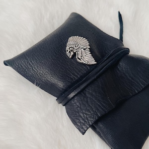 May include: Black leather wrap with a silver-colored Native American headdress pin. The wrap is tied with a black leather cord. The leather has a textured appearance. The pin is detailed with feathers and a skull.