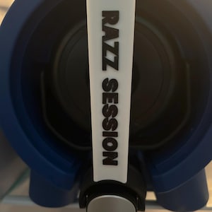 May include: A blue and black beverage dispenser with a white handle. The handle has the words "RAZZ SESSION" printed vertically in black. The dispenser has a silver tap and is likely used for dispensing beverages.