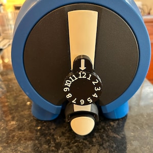 May include: A blue and black device with a white dial and a numbered dial from 1 to 12. The device has a rounded shape with a white rectangular indicator. The device is sitting on a dark surface.
