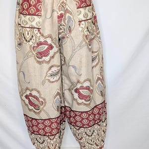 May include: Loose-fitting trousers featuring an elasticized waistband and gathered cuffs at the ankles. The fabric displays a beige base color with a repeating floral and paisley pattern in shades of deep red, muted blue, and dark brown throughout.