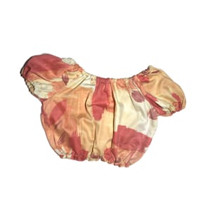 May include: A short-sleeved, off-the-shoulder top with a peach and orange abstract print. The top is made of a soft, stretchy fabric and has a gathered elastic waistband.