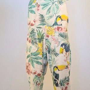 Könnte beinhalten: Weißer Jumpsuit mit tropischem Print mit Tukans, Ananas und floralen Elementen. Der Jumpsuit hat verstellbare Bindeträger und Taschen. Das Design ist eine lebendige Mischung aus Farben.