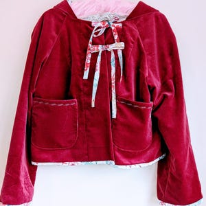 Peut inclure: Une veste en velours bordeaux avec une doublure à imprimé floral. La veste a une capuche et deux poches. La veste est nouée à l'avant avec un ruban floral.